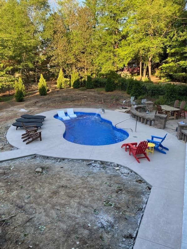 Backyard pool area with a small blue pool, lounge chairs, and colorful patio chairs on a concrete patio.