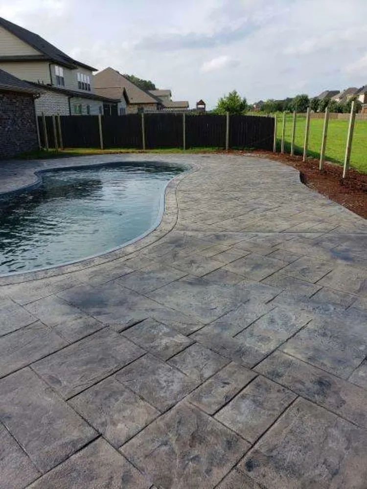 Backyard pool with curved stone patio and black privacy fence under a cloudy sky