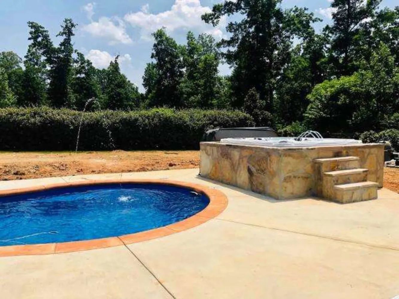 Backyard pool with blue water, tan stone patio, and raised spa with steps under a clear sky