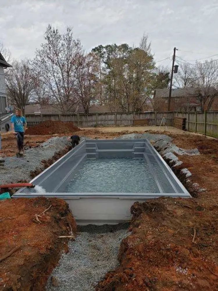 Rectangular concrete pool under construction in a muddy backyard, with water inside and workers nearby
