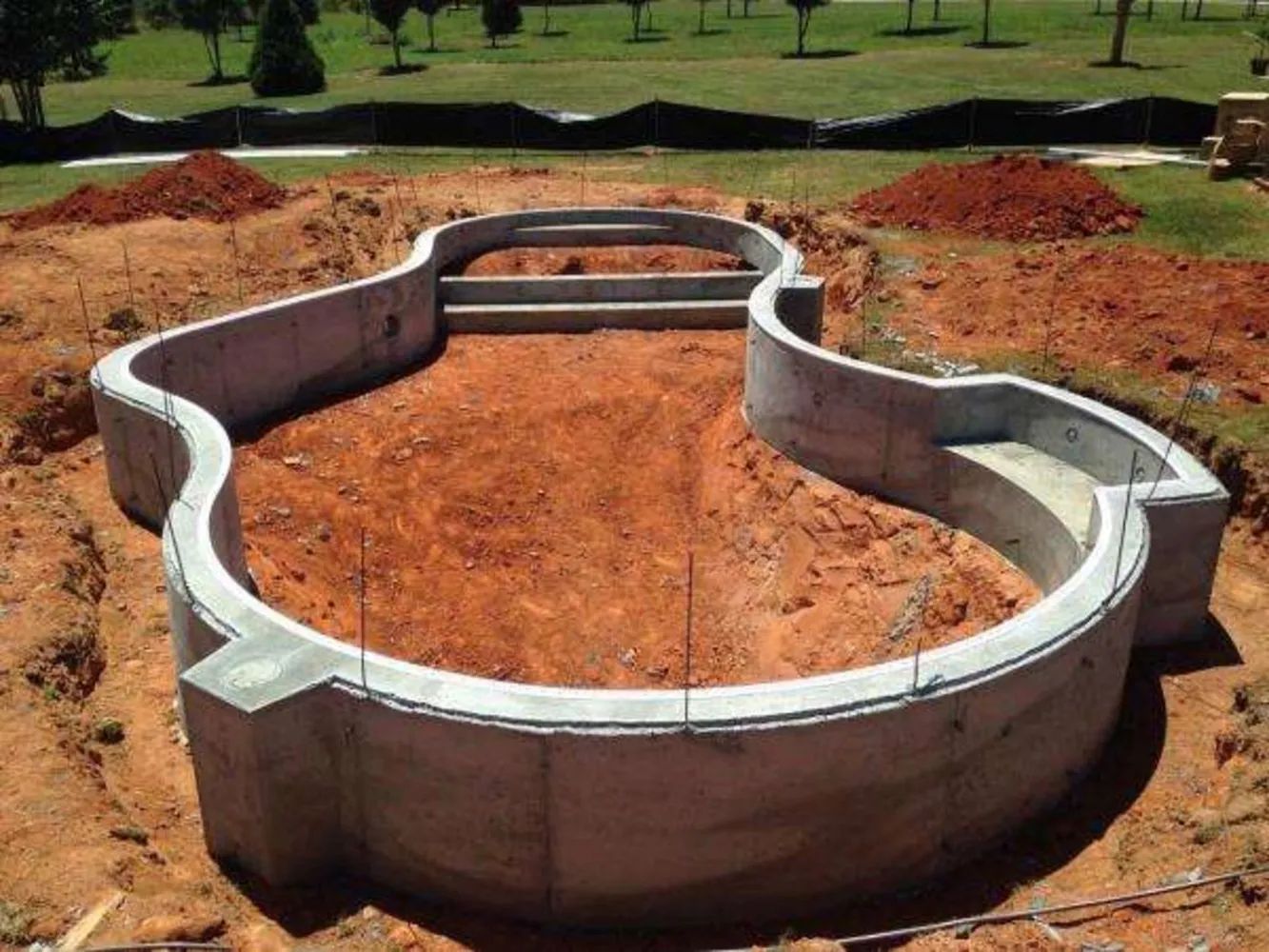 Curving concrete foundation wall under construction on a dirt lot, with red soil mounds around it.