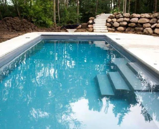 Rectangular blue swimming pool with built-in steps, bordered by white stone and a rock wall