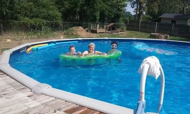 Three people floating in a blue pool on a green raft, with a wooden deck and trees nearby
