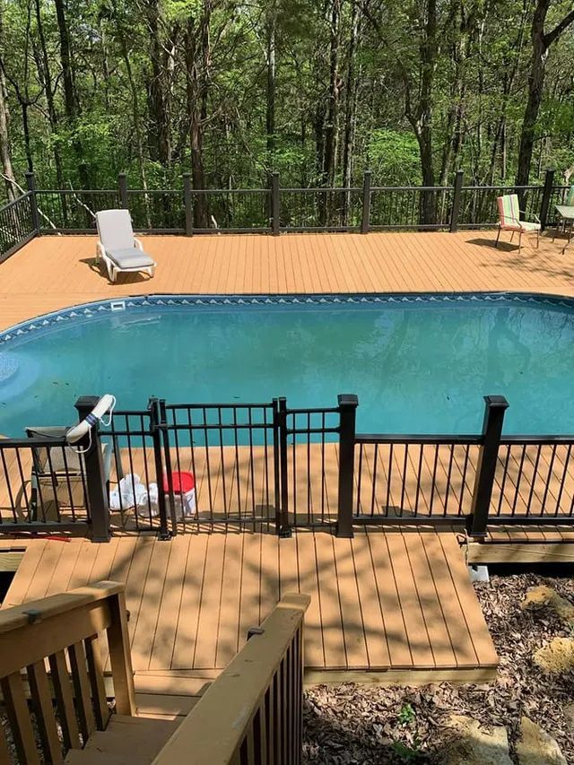 Backyard deck with a swimming pool, black fence, lounge chair, and wooded trees beyond.