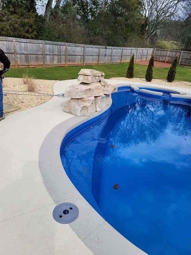 Backyard swimming pool with curved blue water, light deck, rock waterfall, and wooden fence