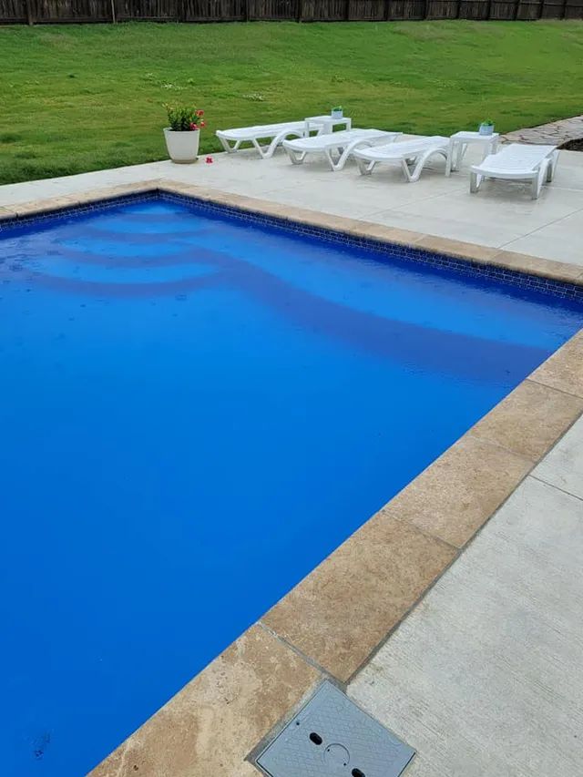 Blue backyard swimming pool with white lounge chairs on a patio and green lawn nearby