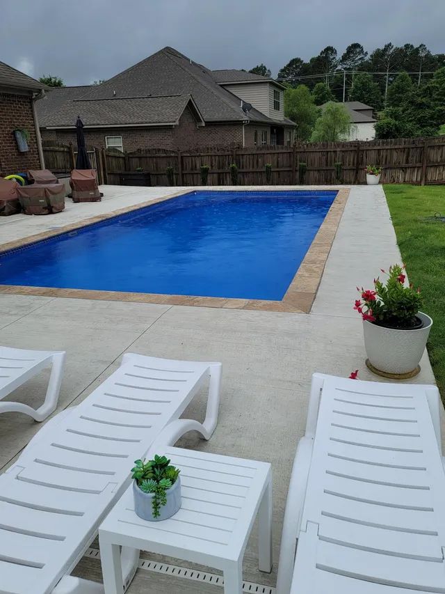 Backyard with blue swimming pool, white patio chairs, and potted flowers under a cloudy sky