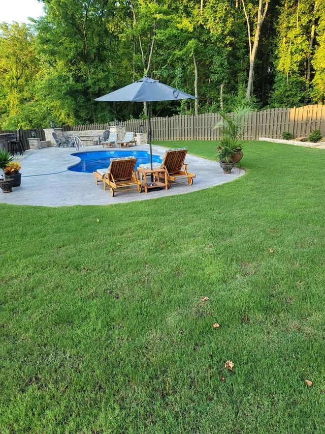Backyard patio with blue umbrella, wooden chairs, and a small pool surrounded by green lawn and trees