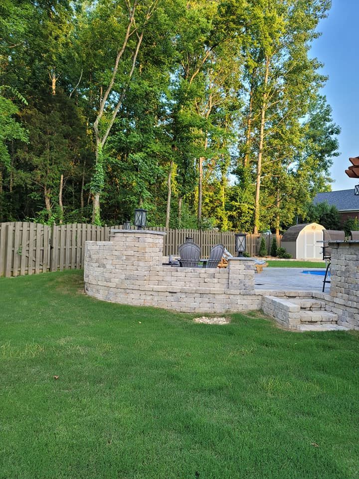 Backyard with green lawn, stone patio and retaining wall, trees, and a small shed in the background