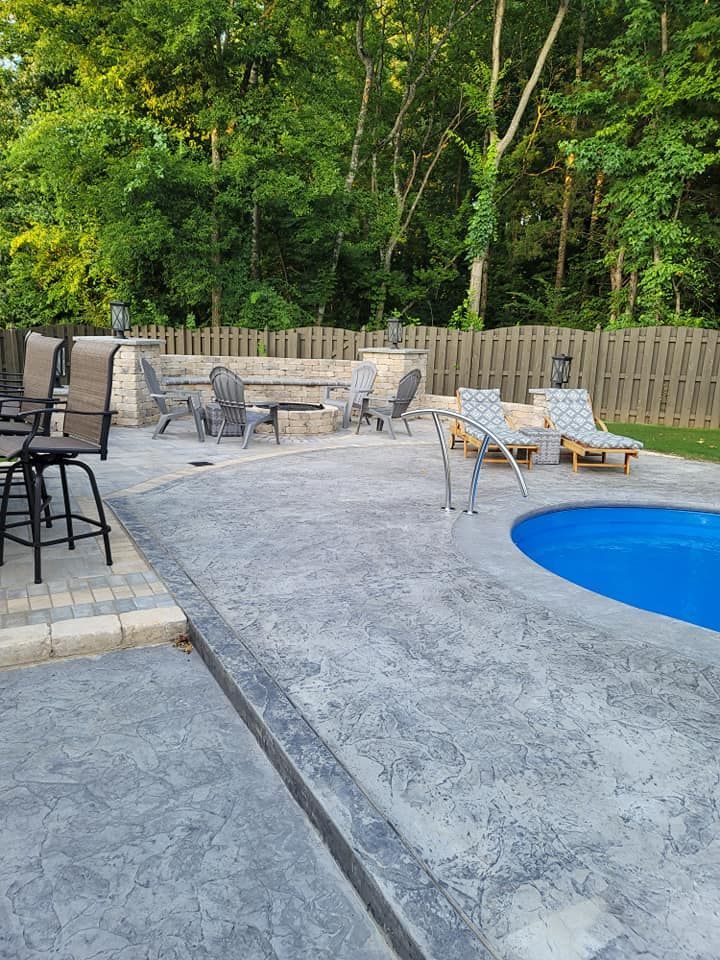Backyard pool deck with patio chairs, table, grill, and a fence backed by trees