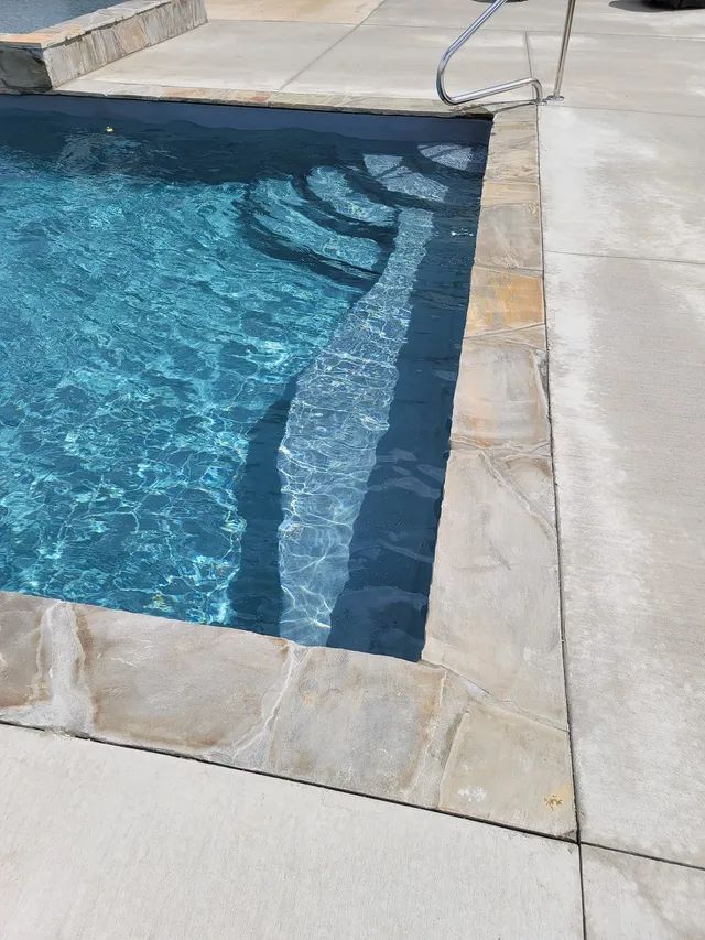 Blue swimming pool with stone coping beside a light concrete deck and steps visible underwater