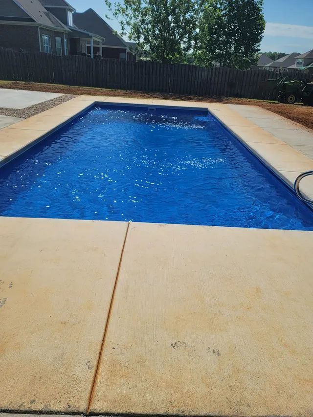 Backyard swimming pool with blue water, beige concrete deck, and a wooden fence in the background