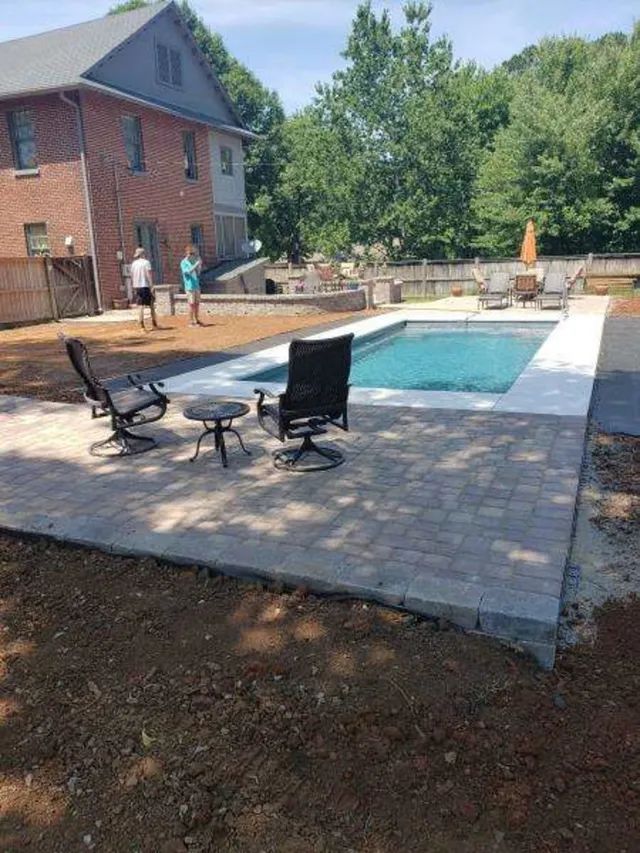 Backyard patio with a swimming pool, lounge chairs, and a brick house beside trees