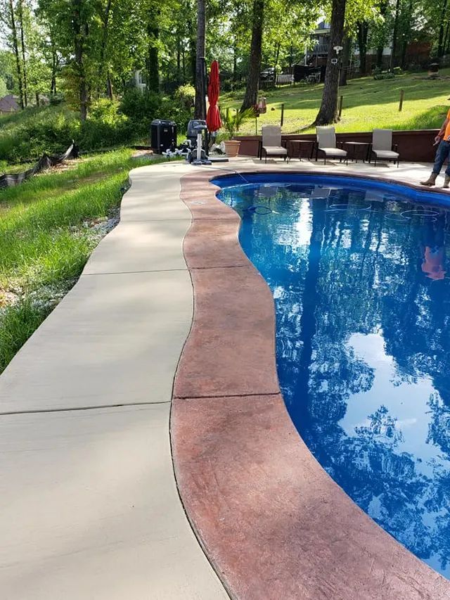 Curved backyard pool with blue water, patio chairs, and trees in a landscaped garden.