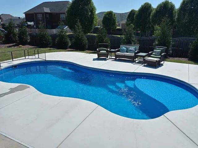 Backyard swimming pool with blue water, patio chairs, and trimmed hedges on a sunny day