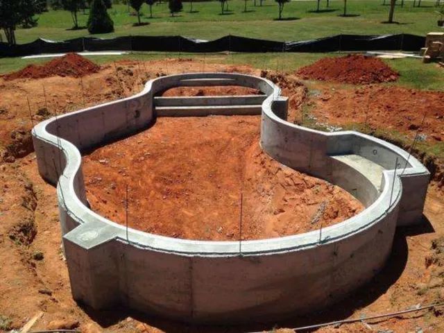 Concrete foundation outline on red dirt at a construction site, surrounded by grassy landscape