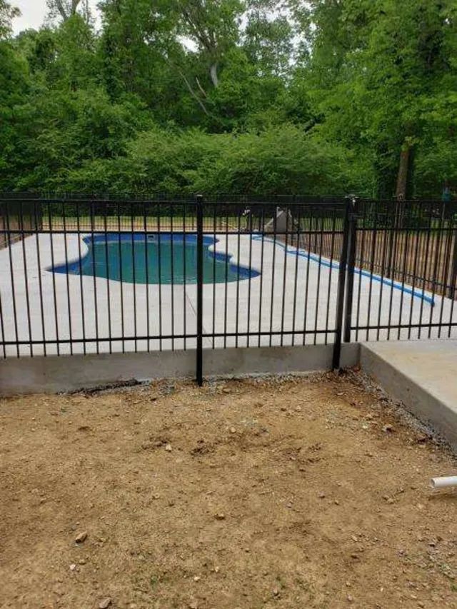 Backyard patio with black fence, tan ground, and covered pool surrounded by trees