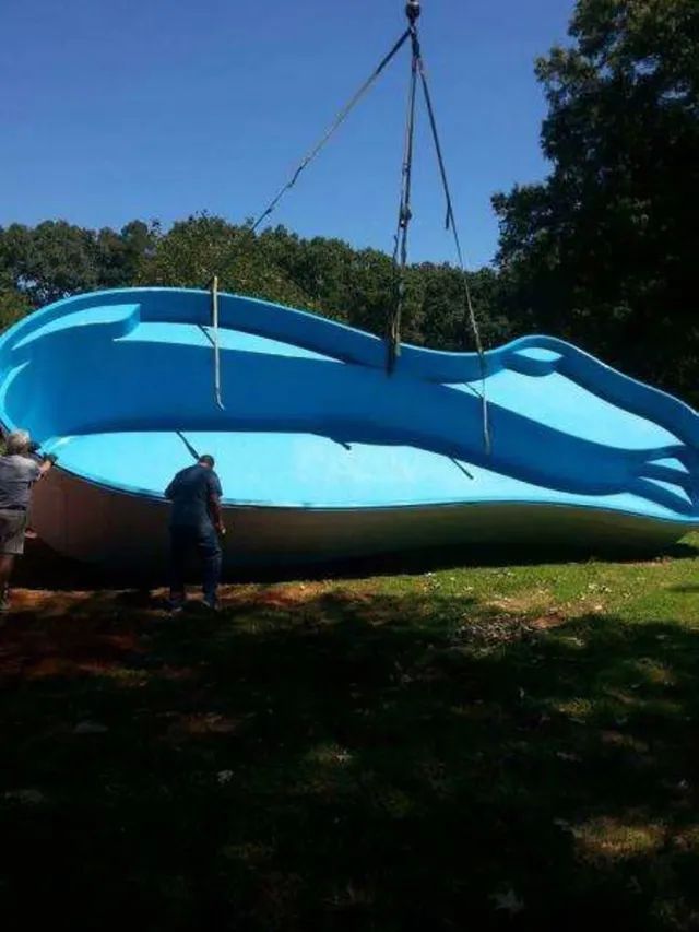 Blue swimming pool shell being lifted by a crane in a grassy yard, with a worker standing nearby.