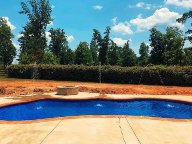 Backyard swimming pool with blue water, tan patio, fountain jets, and trees under a bright sky