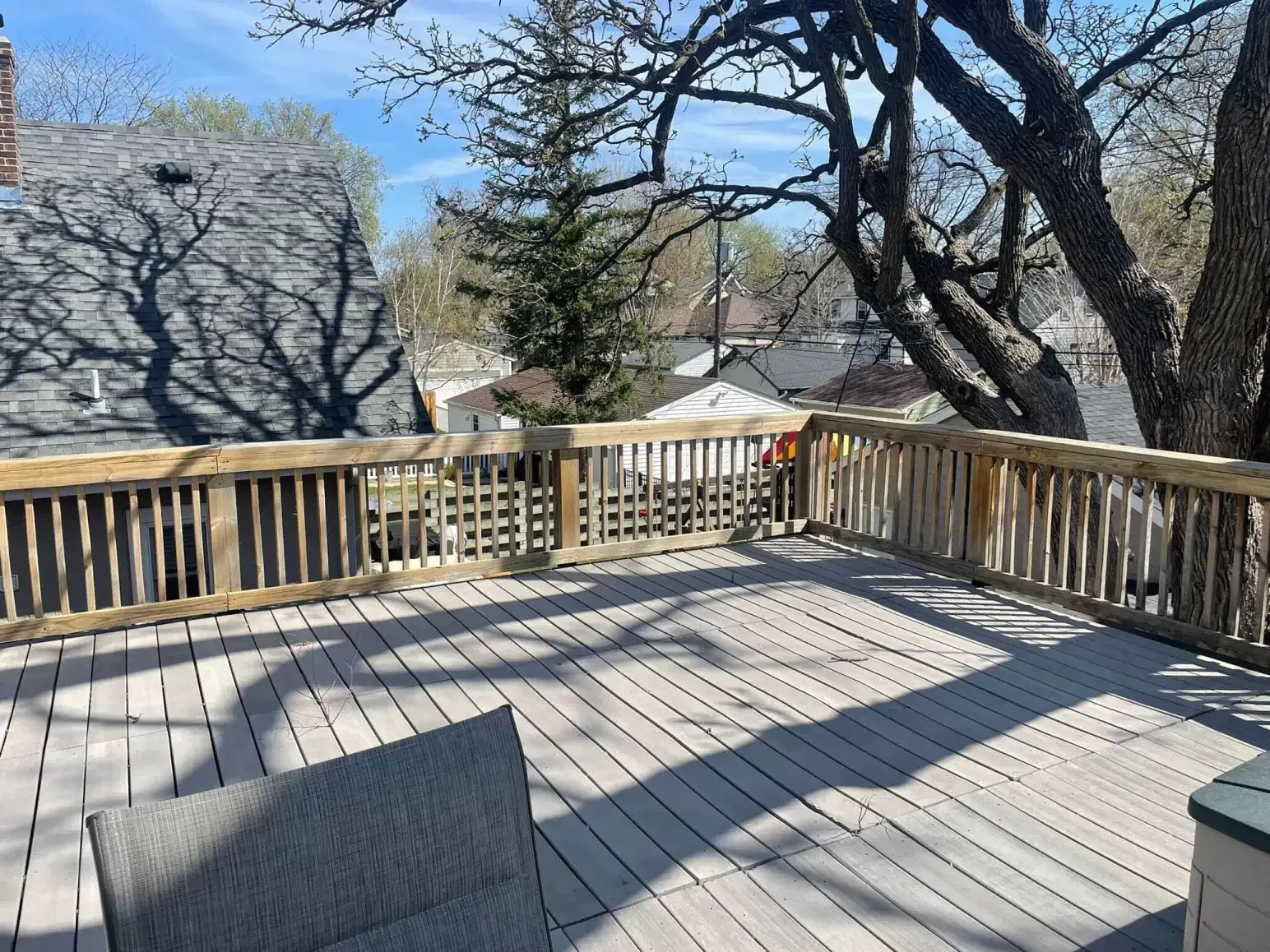 Wooden deck with railing, trees casting shadows, and a glimpse of rooftops in background.