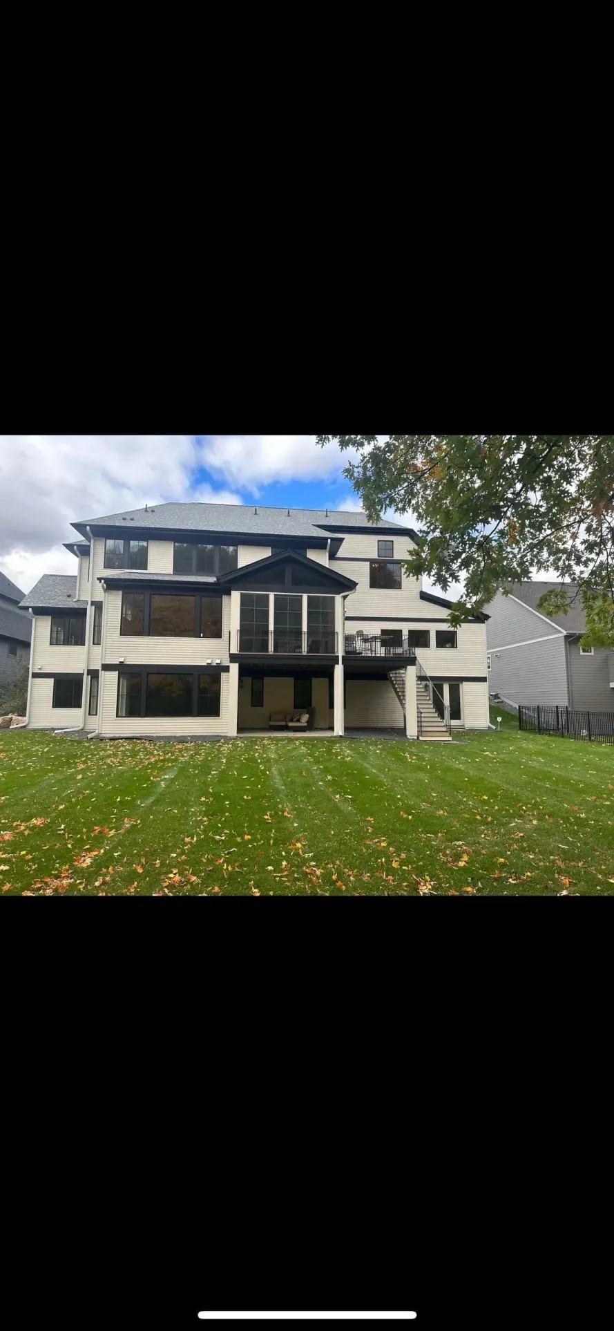 Back of a large, modern home with a green lawn. Cloudy sky, trees on the right, and a black accent above.