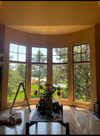 Interior view of large bay windows overlooking a green landscape with a ladder and floral arrangement in the foreground.