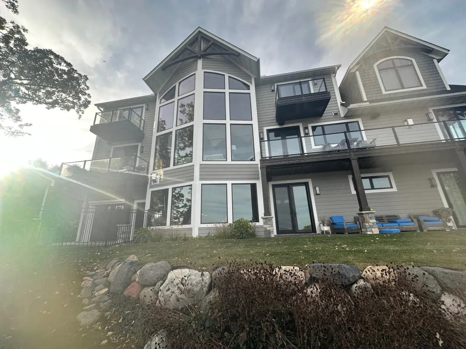 Large gray house with many windows and balconies overlooking a lake, sunny day.