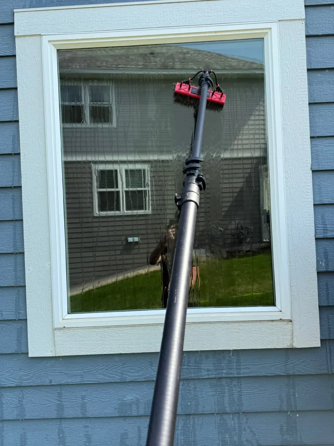 Window being cleaned with a long-handled tool; reflection of house, lawn, and sky.
