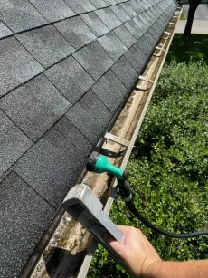 A person cleans a dirty gutter with a hose, near a rooftop.