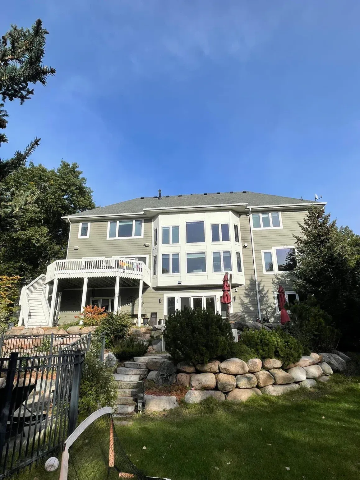Two-story house with green siding, multiple windows, and a deck, under a blue sky. Landscaping with rock wall and green grass.