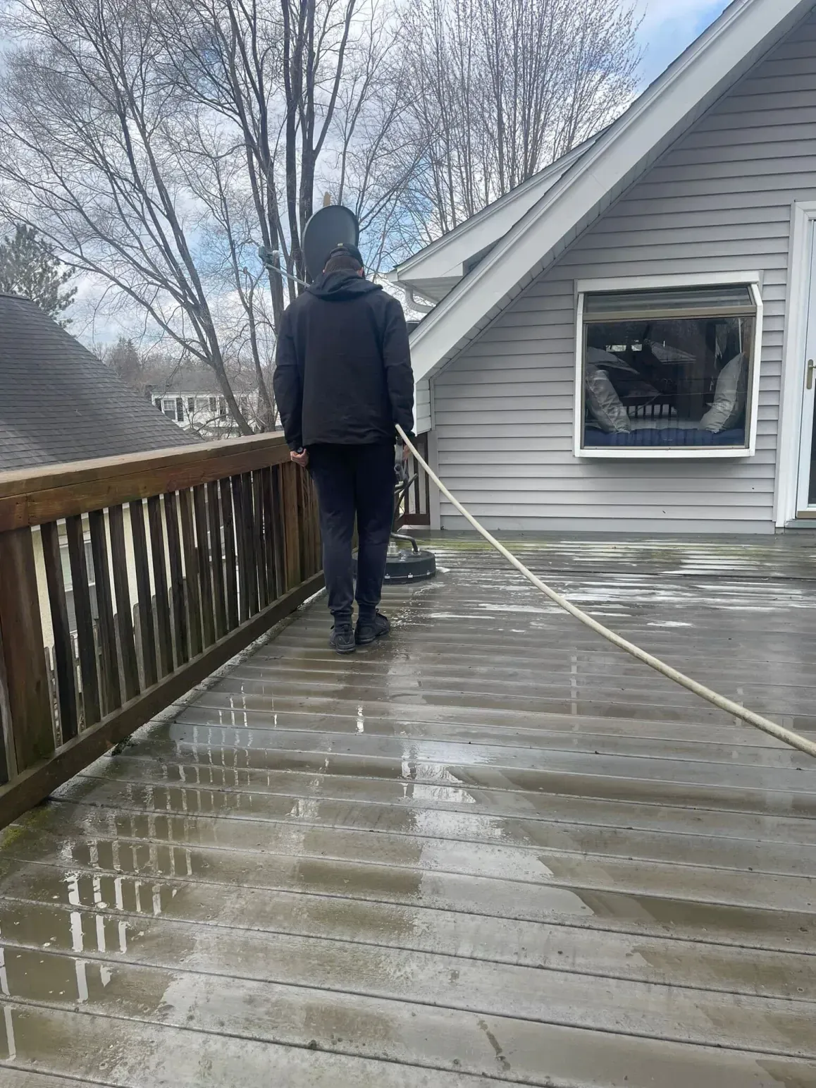 Person pressure washing a wet wooden deck attached to a gray house, on a cloudy day.