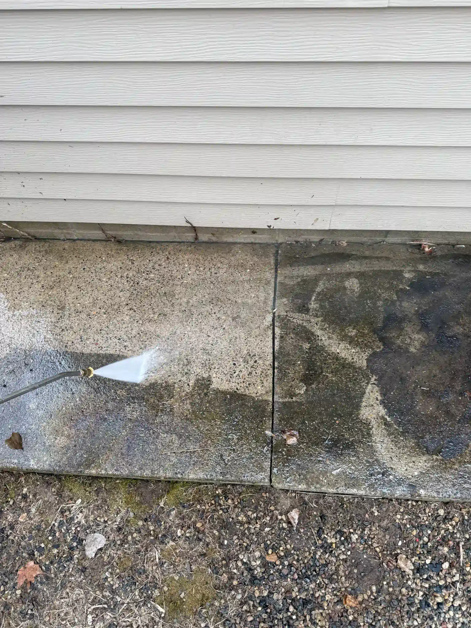 Concrete being power washed. Half is clean, other half is dirty. Gray siding above, gravel below.
