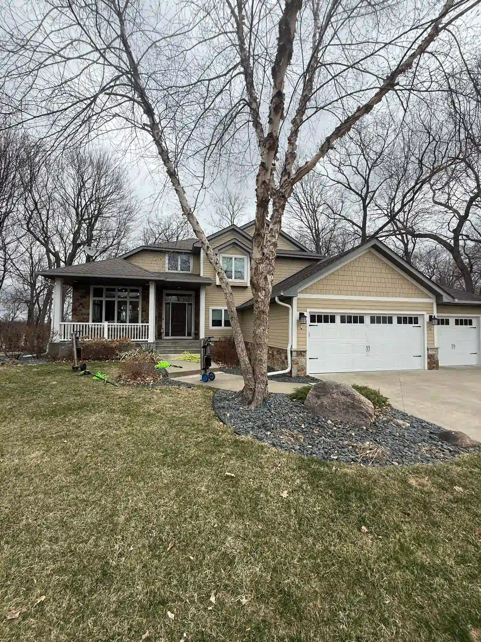 House with beige siding, white garage doors, and a bare tree in front. Overcast day.