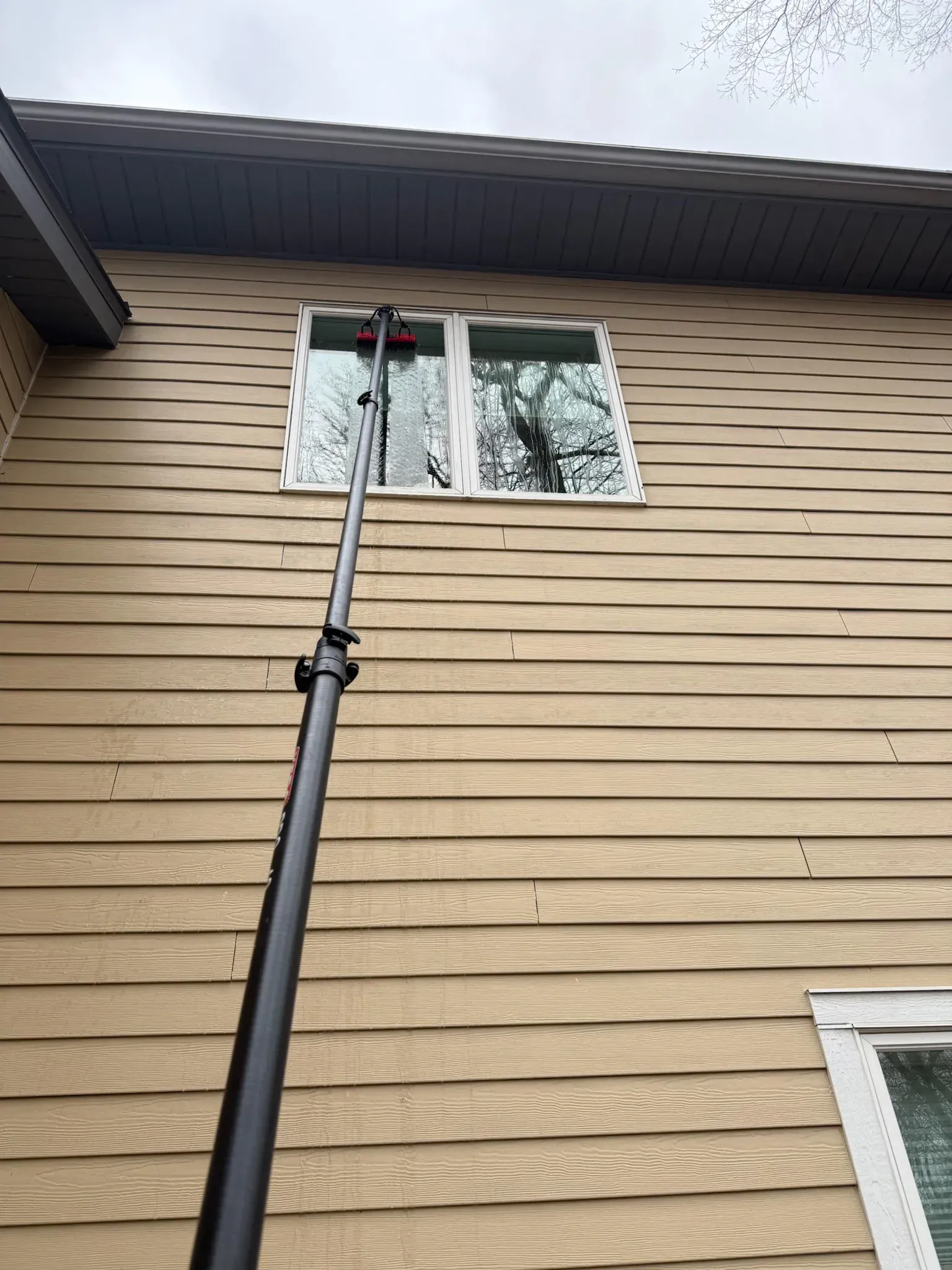 A window being cleaned with a long-handled brush on the side of a tan house.