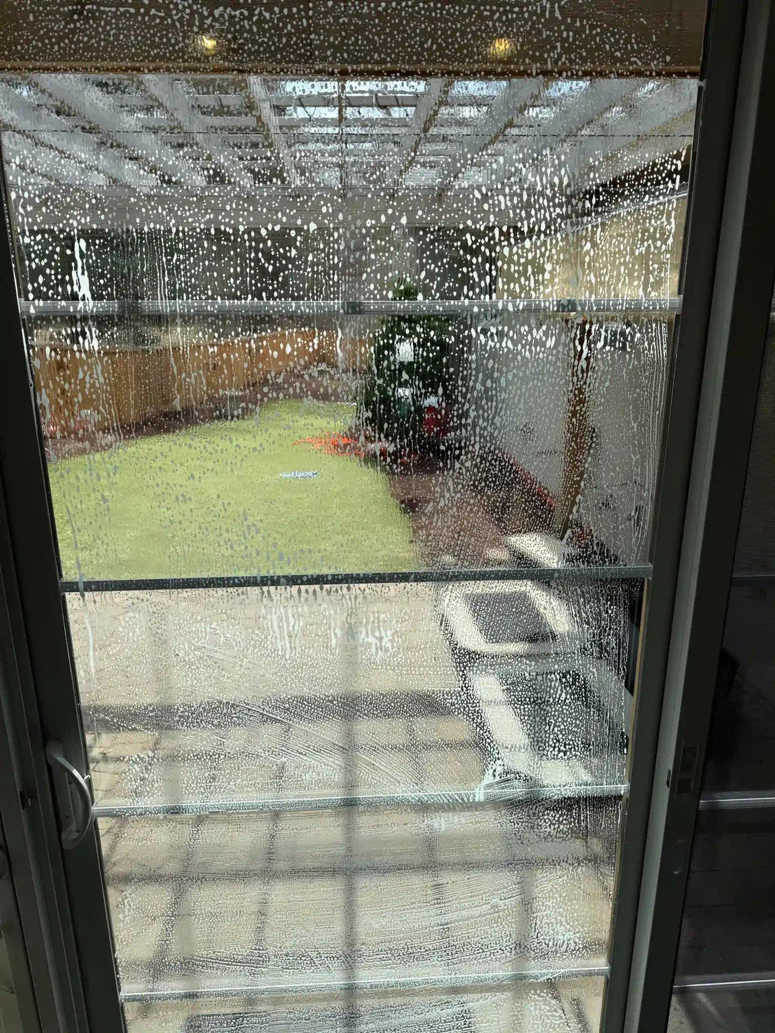View through a rain-streaked window; backyard with green grass, a small tree, and a tiled patio.