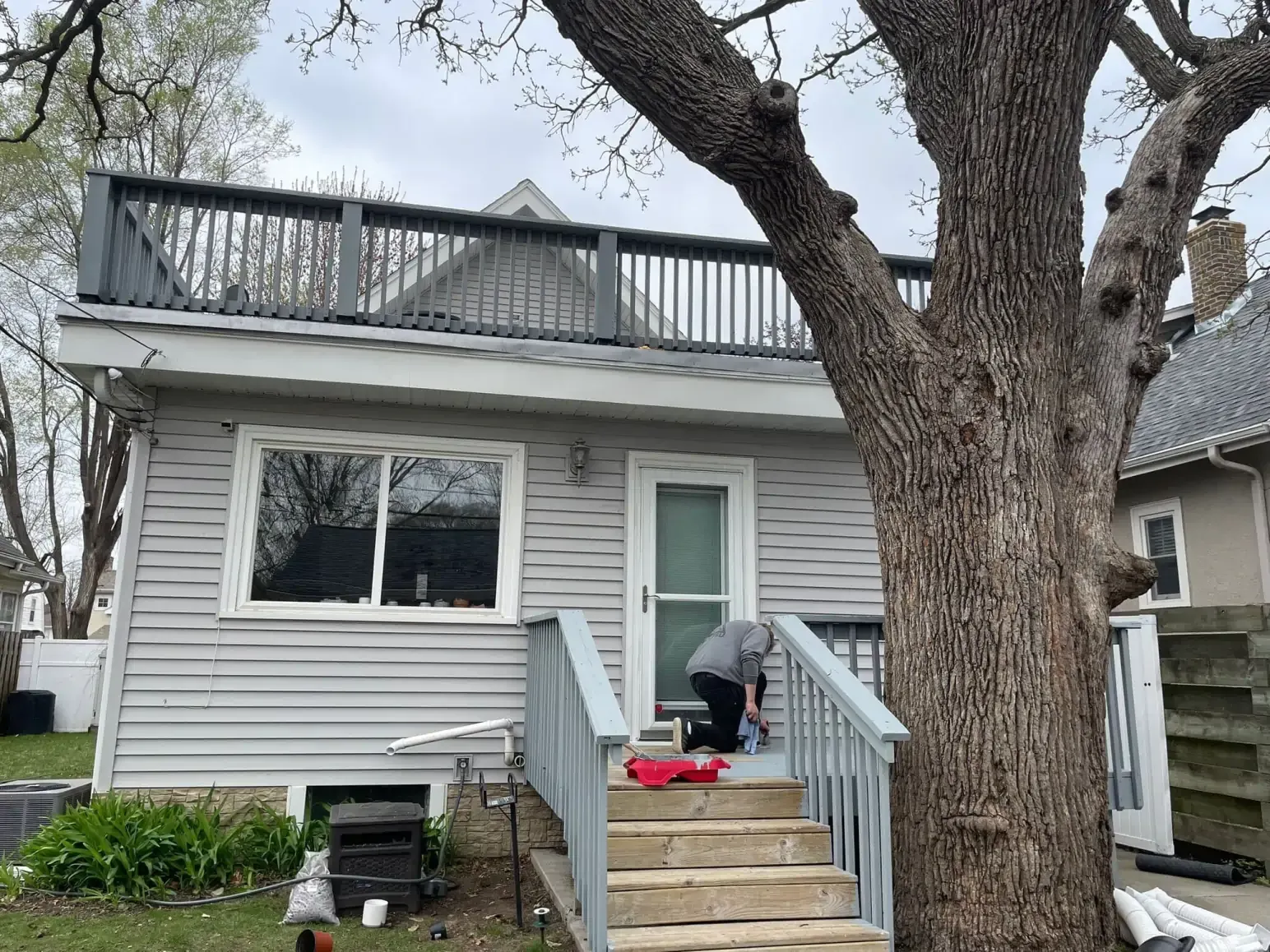 A person on the steps of a light gray house with a rooftop deck under a large tree.