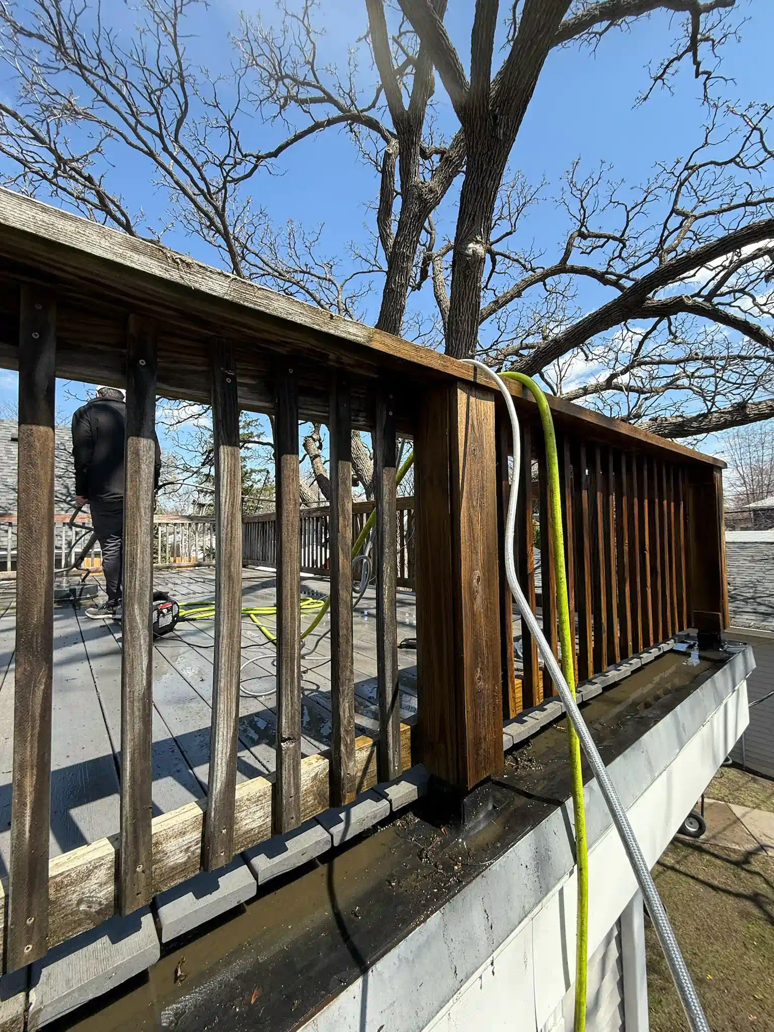 Wooden deck railing being cleaned with a pressure washer; water spray and a blue sky in the background.