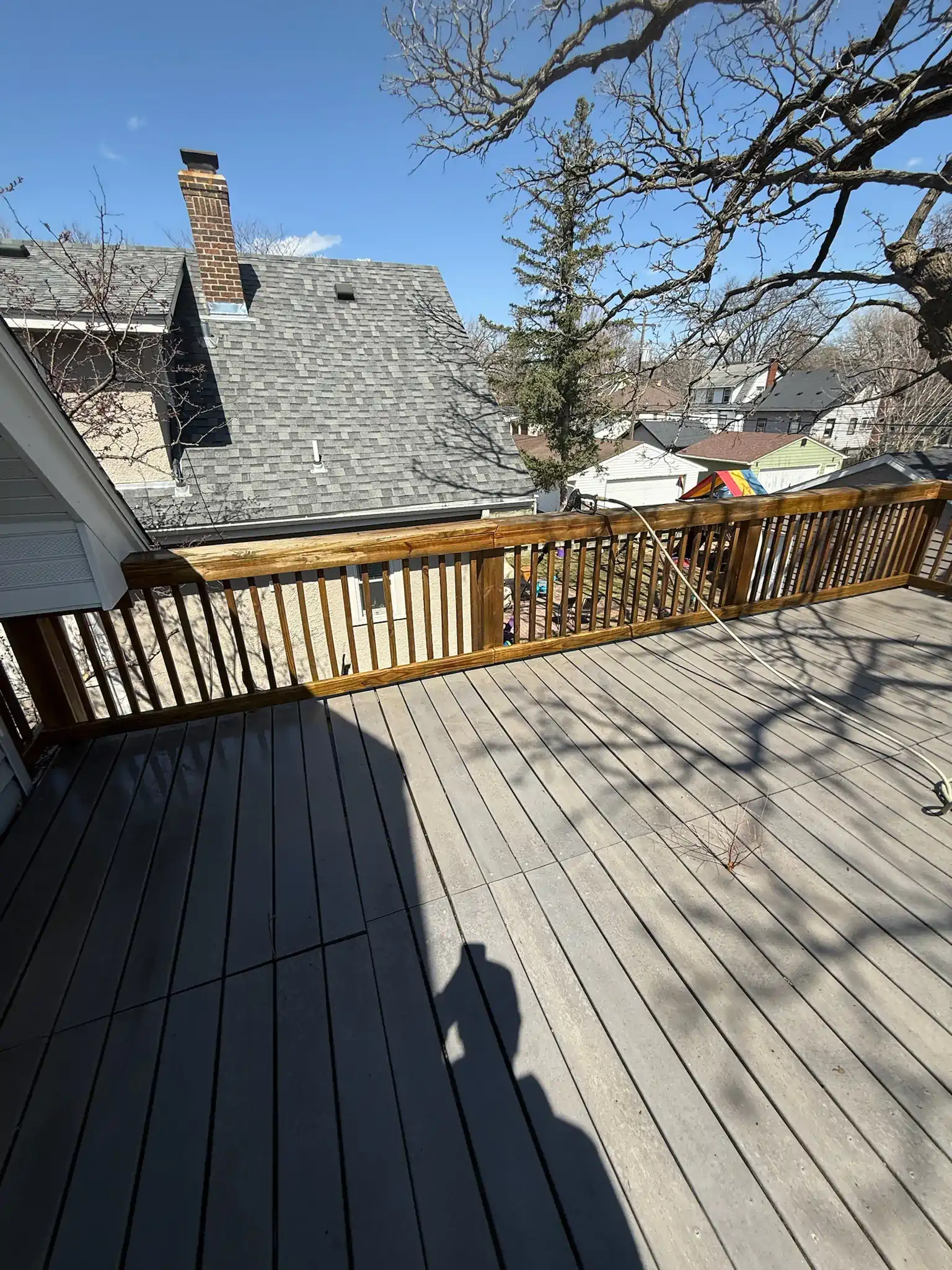 Wooden deck with railing, house roof, and trees under a clear sky.