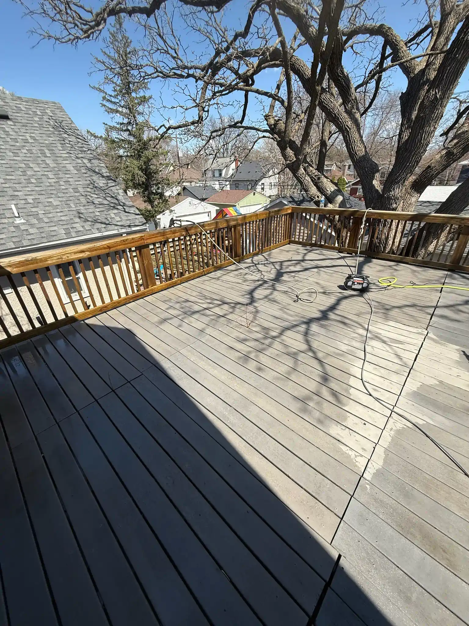Wooden deck with railing, a large tree, and a view of houses on a sunny day.