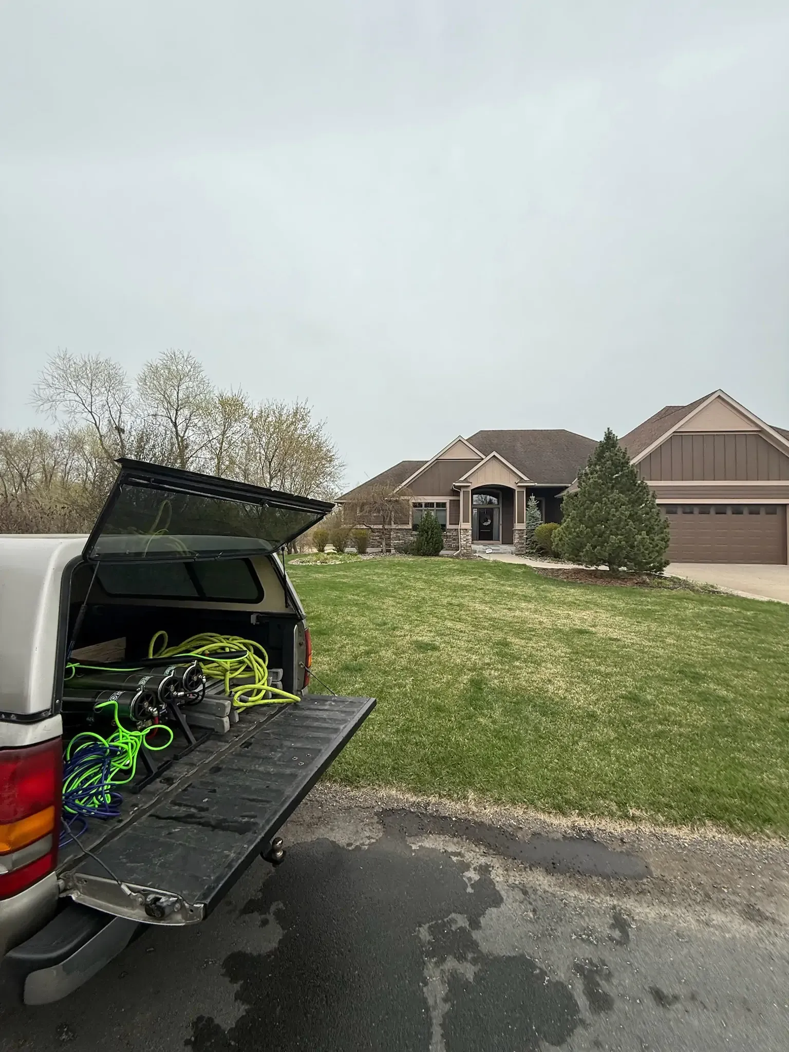 Truck bed open with equipment, facing a house on a grassy lot under a cloudy sky.