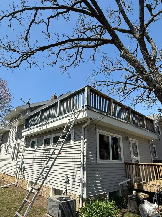 House with a wooden deck, a ladder leading to it, and a leafless tree against a blue sky.