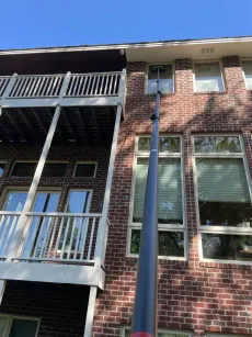 A window cleaner uses a long pole to clean windows on a brick house with a balcony.