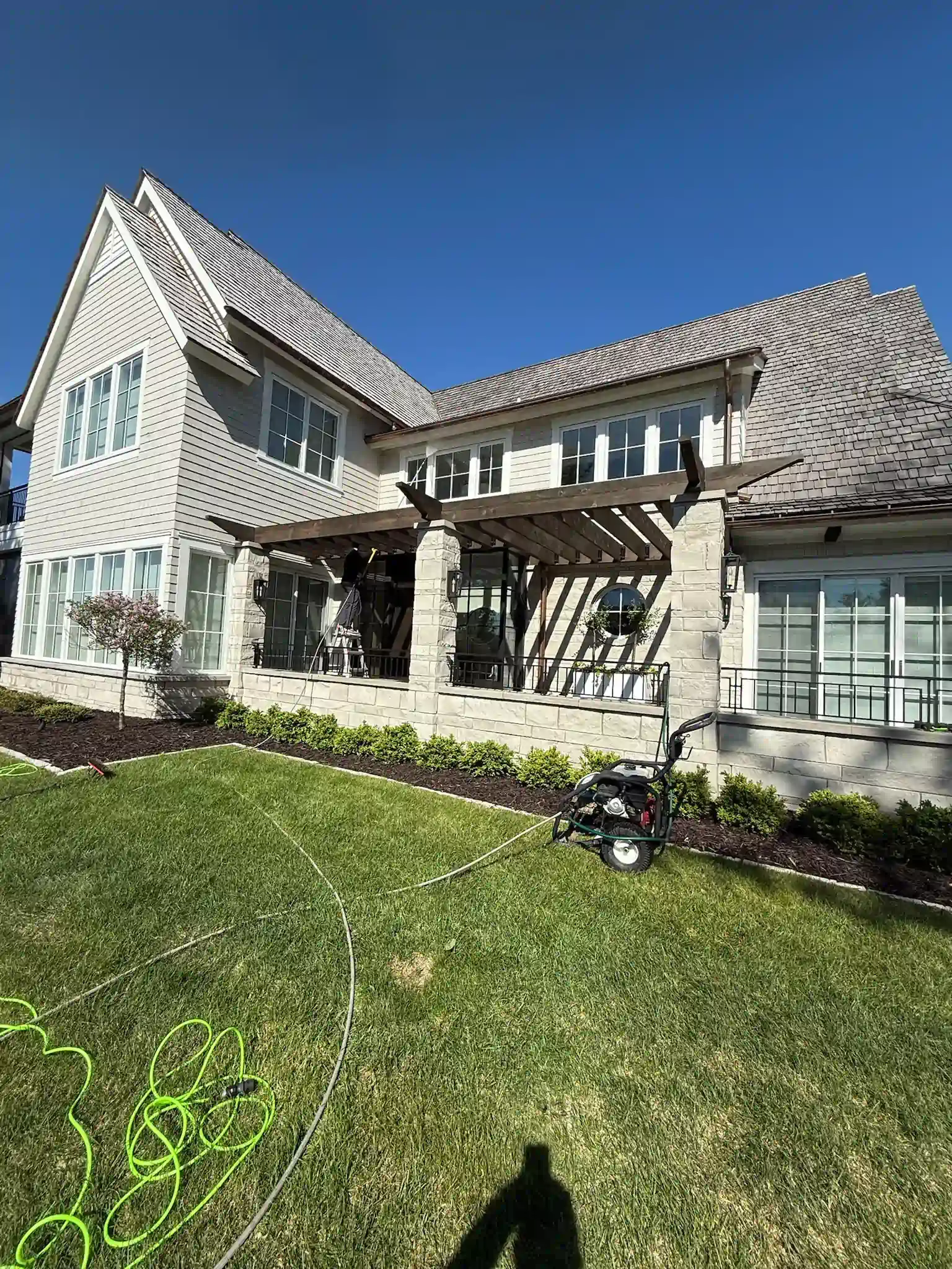 Large two-story house with a pergola over a patio. Green grass and blue sky are visible.