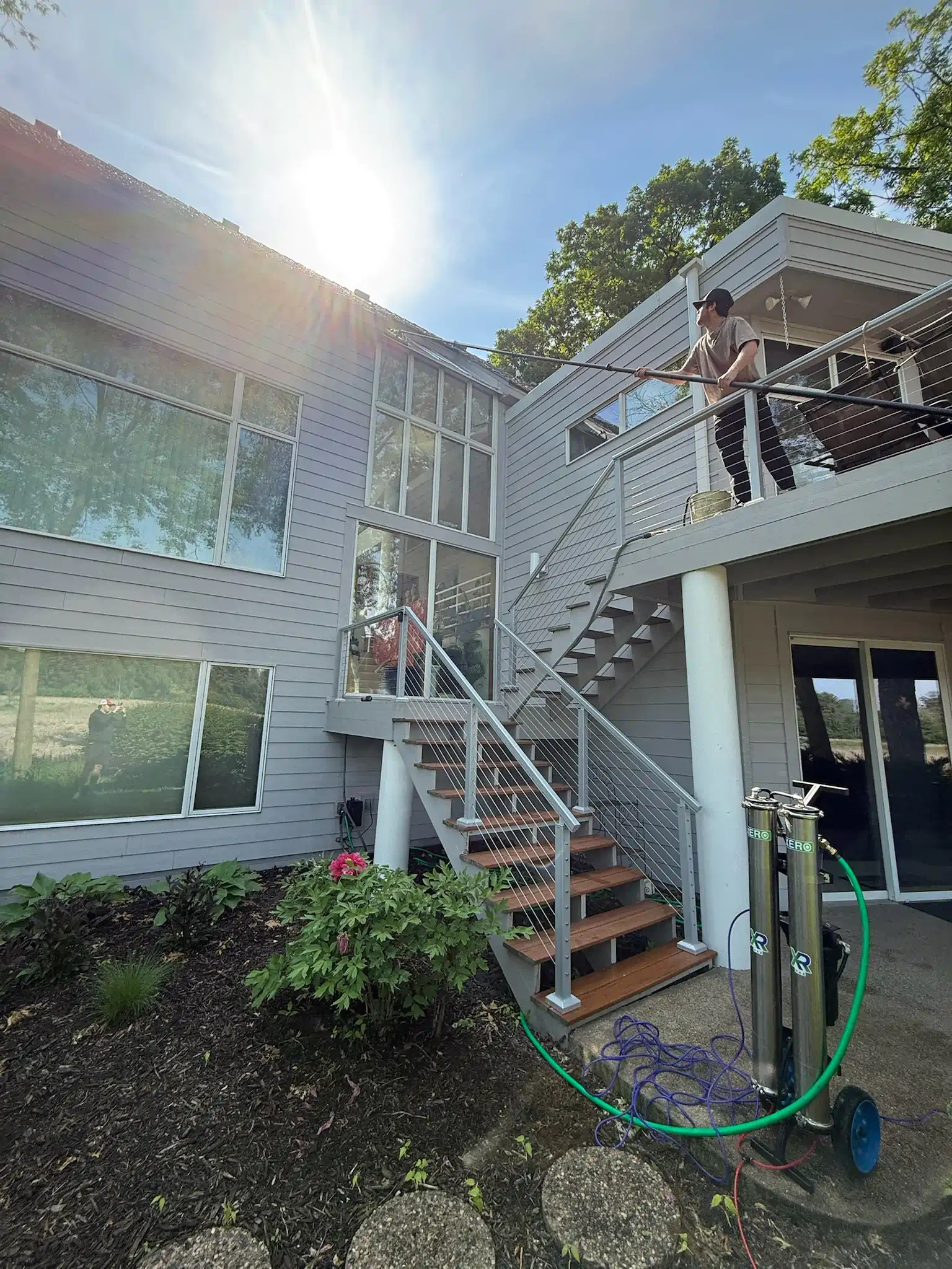 Two-story house with a deck and exterior stairs. Person on deck with hose. Blue sky.
