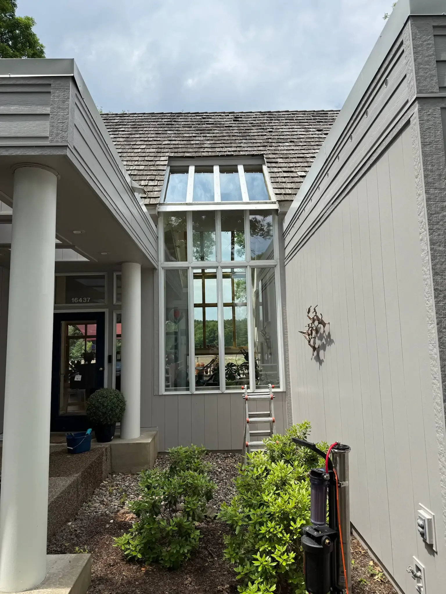 Exterior view of a tall window between two gray walls and a wooden roof, with a ladder and landscaping in front.