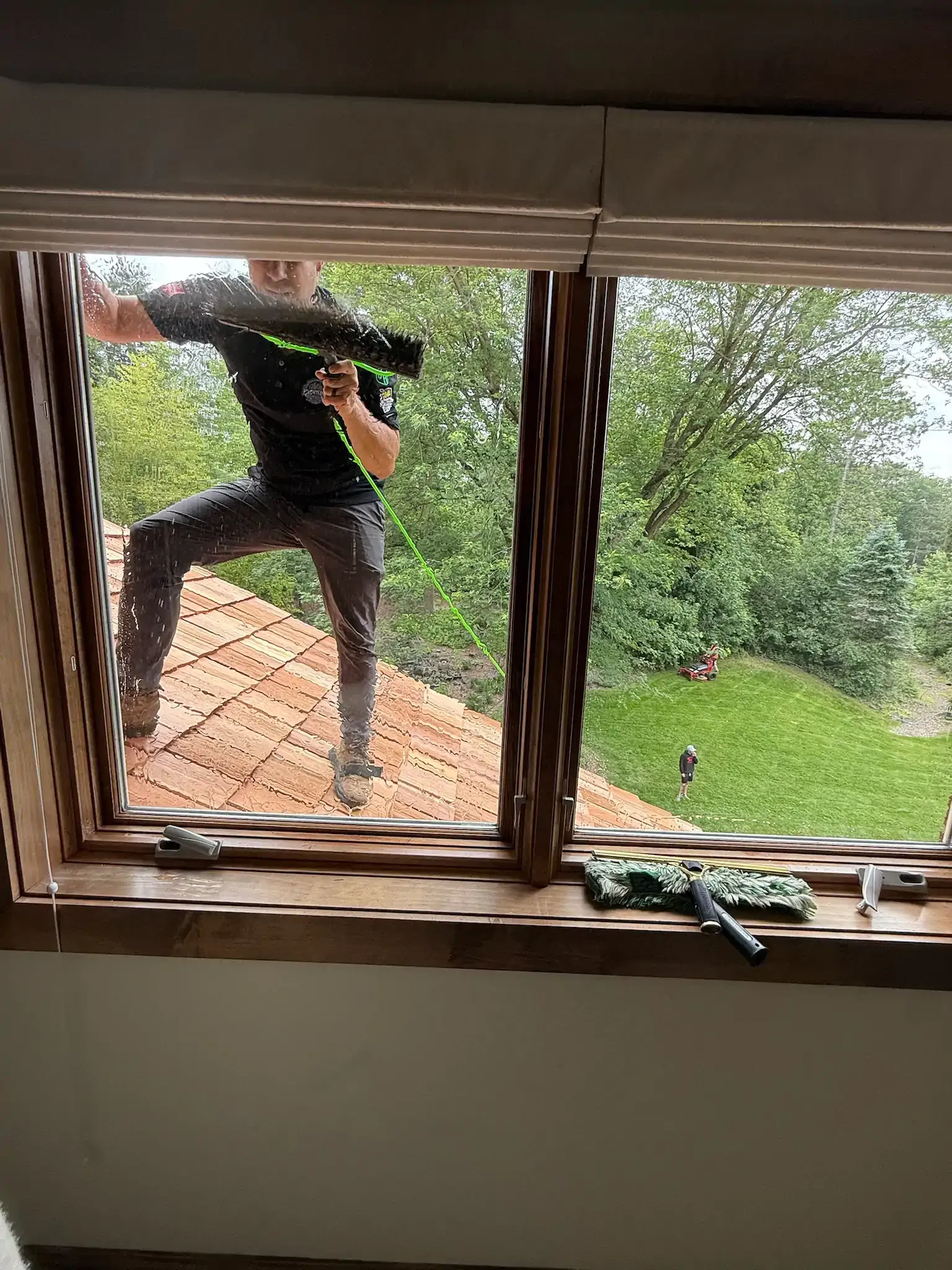 Man cleaning window from outside; greenery and terracotta roof in the background.