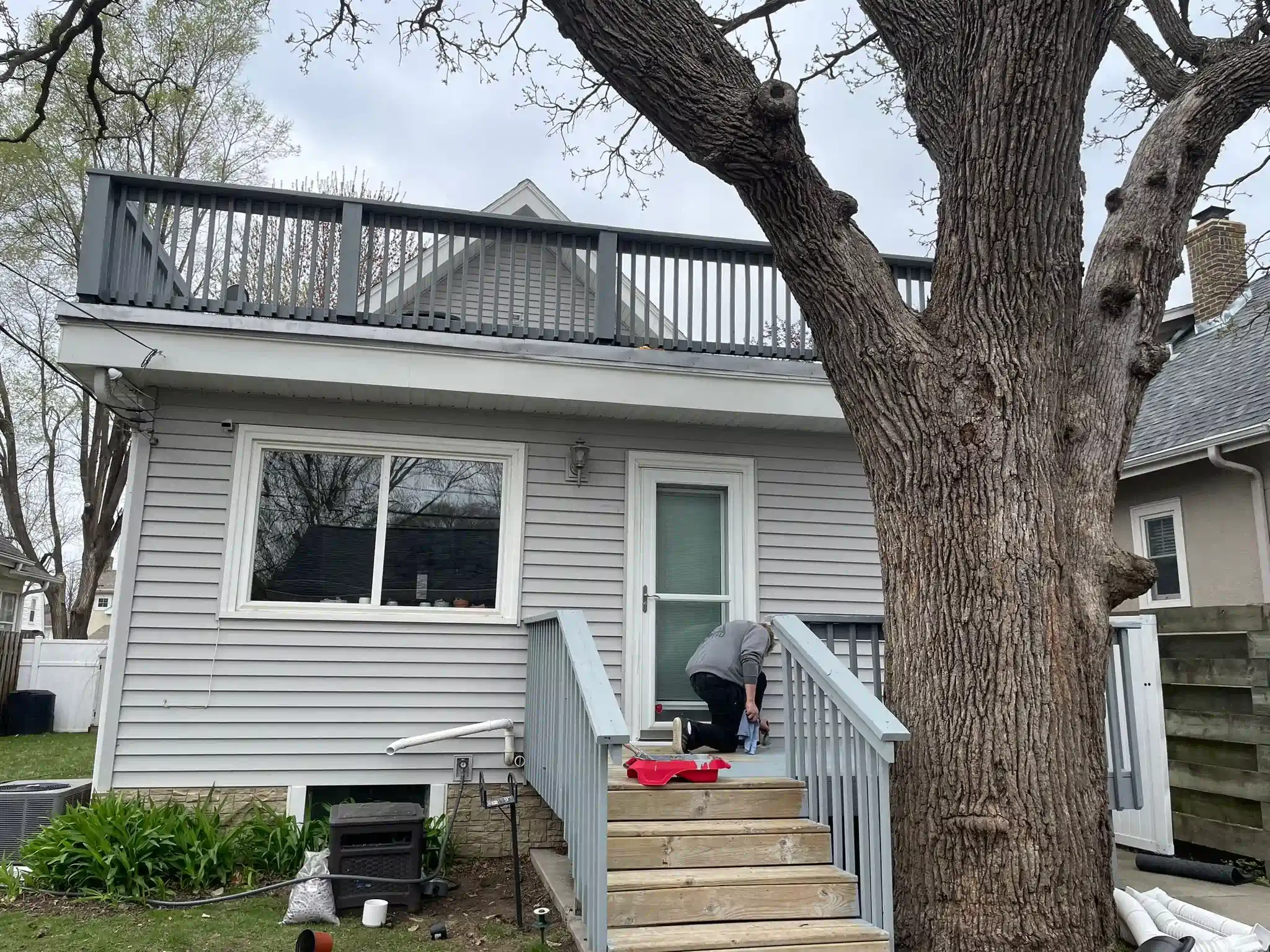 Person on steps near gray house with rooftop deck and large tree.