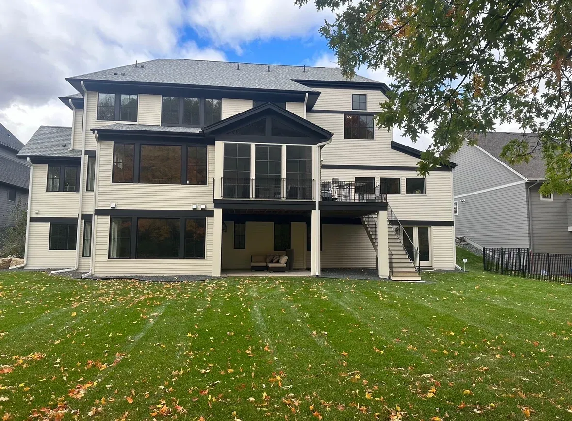 Back of a two-story beige house with black trim and windows, a grass lawn, and a cloudy sky.