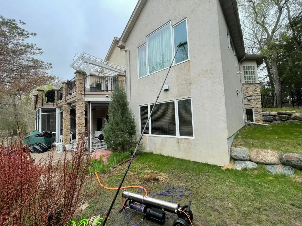 A person washing windows on a two-story beige house with a water-fed pole. Green lawn with bushes and trees.