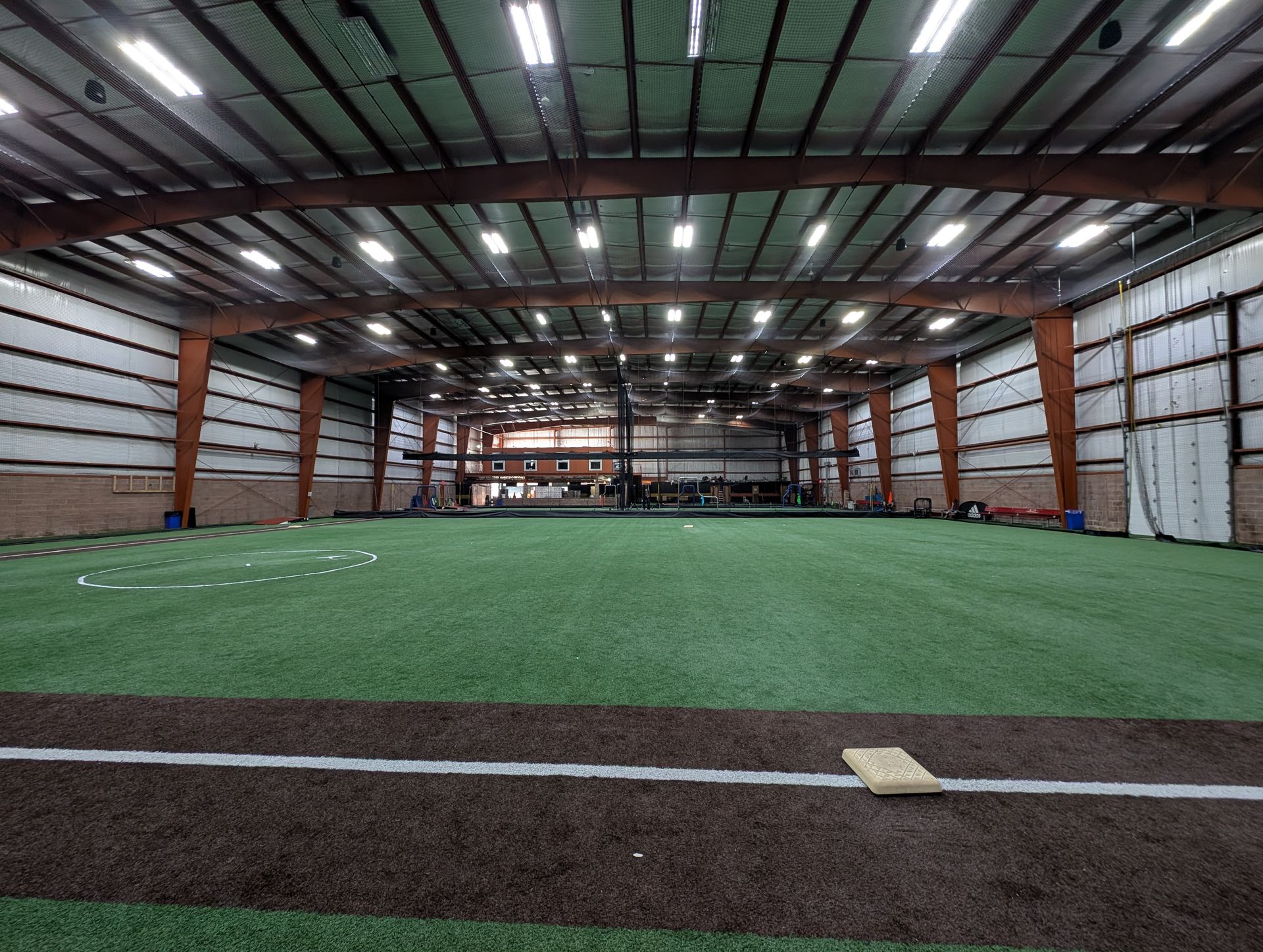 Indoor sports facility with artificial turf, brown and white baseball field markings, and overhead lighting.